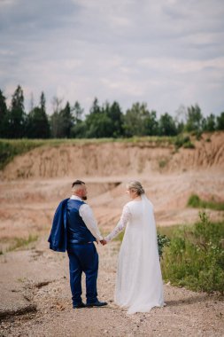 Bir gelin ve damat, manzaralı, kırsal bir arazide kayalık bir yolda dururken el ele tutuşuyorlar. Damat ceketini tutuyor ve birbirlerine sevgiyle bakıyorlar..