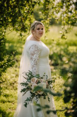 Beyaz dantelli bir gelinlik içinde bir buket tutan ve omuzlarının üzerinden bakan yeşil ve yumuşak doğal ışıkla çevrili güneşli bir bahçede duran bir gelin..