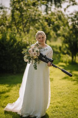 Beyaz dantelli bir gelinlik giymiş bir gelin elinde bir buket ve tüfekle dışarıda duruyor. Dekor güneşli yeşil bir bahçe, zarafeti cesur, eşsiz bir kıvrımla harmanlıyor..