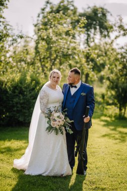 Bir gelin ve damat düğün günlerinde dışarıda dururlar. Damat tüfeği tutarken gelin buketi tutuyor. Güneşin altında birlikte poz verirler..