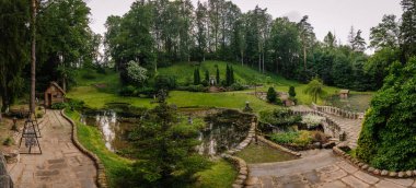 Cesis, Letonya - 17 Temmuz 2025 Svetavots 'un panoramik manzarası göletleri, taş yolları, heykelleri, ağaçları ve yemyeşil yeşilliklerle çevrili küçük ahşap bir evi olan güzel bir bahçe.