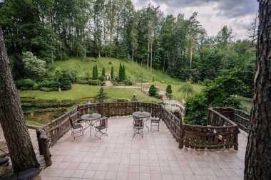 Cesis, Letonya - 17 Temmuz 2025 - Svetavots, Letonya 'da heykeli, göleti, taş basamakları ve yemyeşil bir ormanı olan bahçeye bakan demir masaları ve sandalyeleri olan sakin bir açık teras.