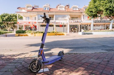 Kemer, Turkey - September 19, 2025 - A parked purple Beam electric scooter stands on a city sidewalk in front of small shops and trees on a bright sunny day in a quiet urban area.