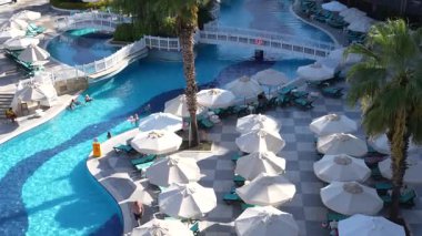 Kemer, Turkey - September 19, 2025 - Luxurious resort pool area with sun loungers, white umbrellas, palm trees, and a winding pool connected by bridges. Guests relax and swim under the sunny sky.