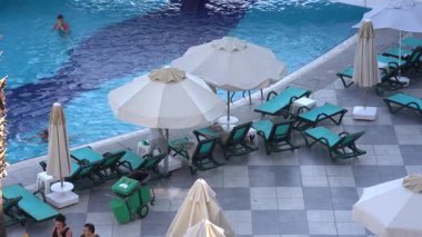 Kemer, Turkey - September 19, 2025 - Outdoor swimming pool with turquoise lounge chairs, beige umbrellas, and a few people relaxing and swimming; poolside scene at a resort hotel.