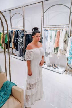 Woman in a white off-shoulder lace dress stands in a modern clothing store, surrounded by fashion racks and shoes. Bright, clean retail environment.