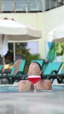 A woman emerges from water in a pool, swims to a red cocktail in a glass, picks it up, and turns away, enjoying a relaxed resort moment.