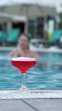 A woman emerges from water in a pool, swims to a red cocktail in a glass, picks it up, and turns away, enjoying a relaxed resort moment.