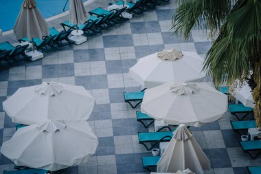 Beyaz şemsiyeli, turkuaz güneş şezlonglu, fayanslı döşemeli ve palmiye yapraklı havuz kenarı manzarası rahatlatıcı bir yaz tatili havası yaratıyor..