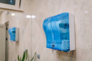 Umumi bir tuvalette duvara monte edilmiş mavi tuvalet kağıdı, fayanslı bir duvara yansıyor, ticari tuvaletlerde hijyen ve rahatlık için kullanılıyor..