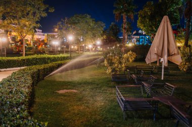Kemer, Turkey - September 21, 2025 - Night view of a beautifully lit park or resort garden with a sprinkler watering the grass, benches, and lush greenery under glowing street lamps.