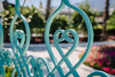 Close-up of a turquoise wrought iron fence with a heart-shaped design, with a blurred background of flowers, trees, and a sunny outdoor garden.