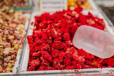 A vibrant pile of dried red flowers displayed in a market bin, with a plastic scoop placed on top, likely used for tea, potpourri, or decoration.