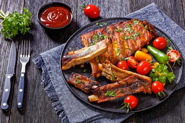 Oven Baked Ribs with fresh tomatoes, chili peppers on black platter on dark wooden table, horizontal view from above