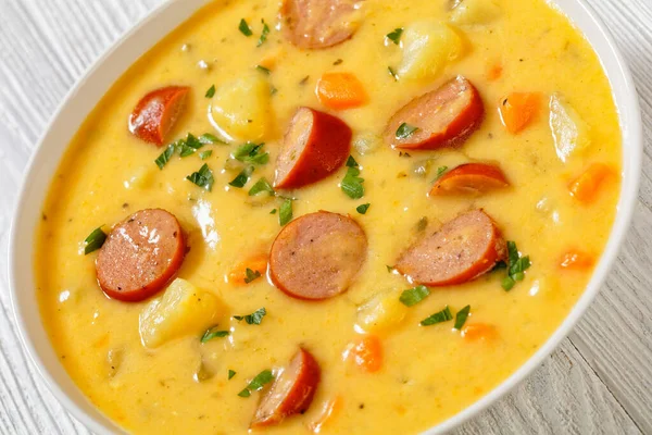 Kartoffelsuppe, German creamy potato soup with sausages in white bowl on white wooden table, close-up