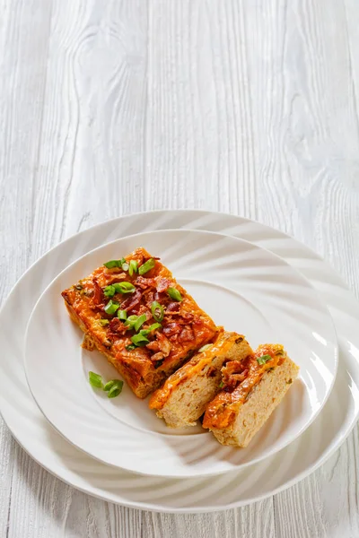 Riced Cauliflower Breakfast Bake topped with melted cheese, green onion and crispy fried bacon on white plate on white wooden table, vertical view, free space
