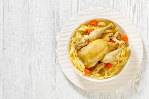 hot chicken soup of whole chicken, egg noodles, celery, onion and carrot in white bowl on wood table, flat lay, free space