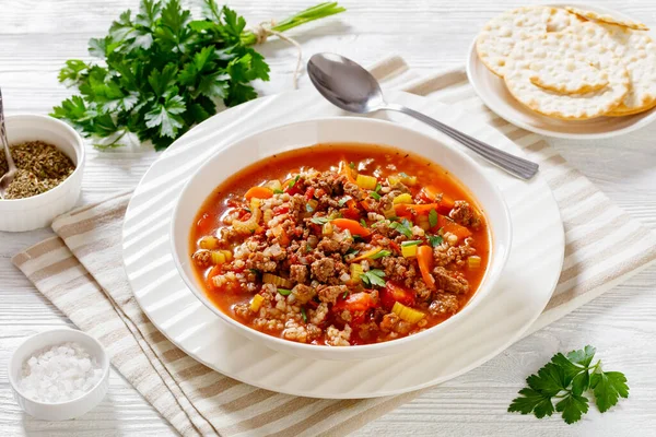 Easy hot and hearty Hamburger Soup with barley and vegetables in white bowl on white wood table, horizontal view from above, close-up