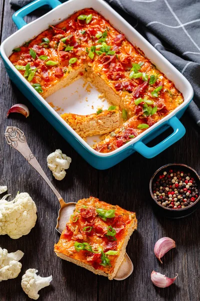 Riced Cauliflower Breakfast Bake topped with melted cheese, green onion and crispy fried bacon in baking dish on dark wooden table, vertical view