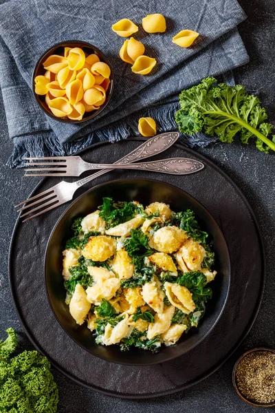 portion of cheesy baked pasta with green leafy kale topped with crispy breadcrumbs in black bowl on concrete table with ingredients, vertical view from above