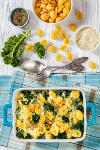 cheesy baked shell pasta with green leafy kale topped with crispy breadcrumbs in baking dish with ingredients on white wood table, vertical view