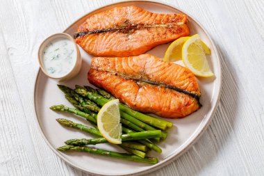 roast salmon steaks with boiled asparagus, fresh lemon and yogurt dill sauce on  plate on white wooden table clipart
