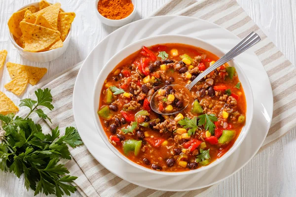 close-up of taco soup of ground beef, tomatoes, chopped green chilis, onions, corn, red beans and taco seasoning in white bowl, horizontal view from above