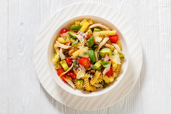 Healthy chicken pasta salad with avocado, tomato, olive oil and vinegar dressing in a black bowl on a dark wood table, horizontal top view, flat lay, free space