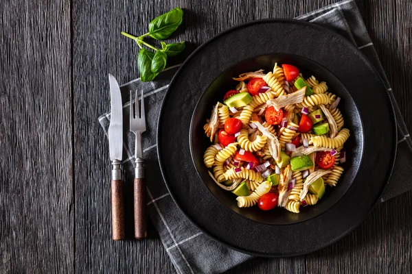 Healthy chicken pasta salad with avocado, tomato, olive oil and vinegar dressing in a black bowl on a dark wood table, horizontal top view, flat lay, free space