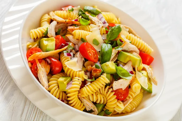 close-up of Healthy Chicken Pasta Salad with Avocado, Tomato, and olive oil and vinegar dressing in white bowl on white wood table,  horizontal view from above