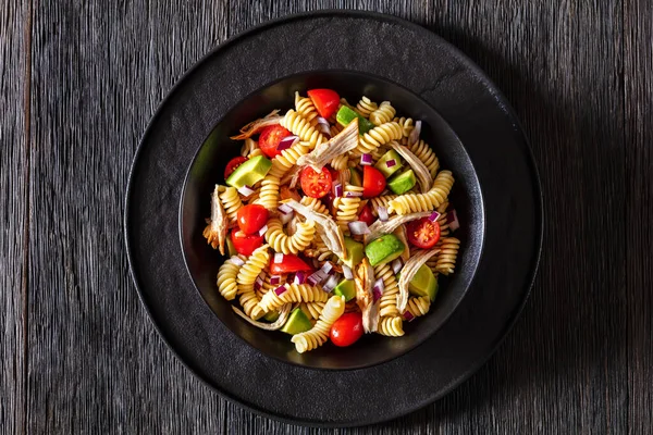 Healthy chicken pasta salad with avocado, tomato, olive oil and vinegar dressing in a black bowl on a dark wood table, horizontal top view, flat lay, closeup