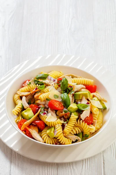 Healthy Chicken Pasta Salad with Avocado, Tomato, and olive oil and vinegar dressing in white bowl on white wood table,  vertical view from above, free space