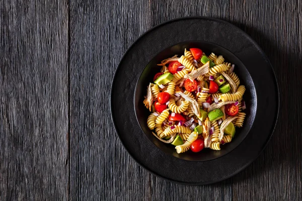 Healthy chicken pasta salad with avocado, tomato, olive oil and vinegar dressing in a black bowl on a dark wood table, horizontal top view, flat lay, copy space