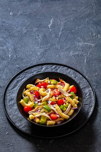 Healthy Chicken Pasta Salad with Avocado, Tomato, and olive oil and vinegar dressing in black bowl on table, vertical view from above, free space