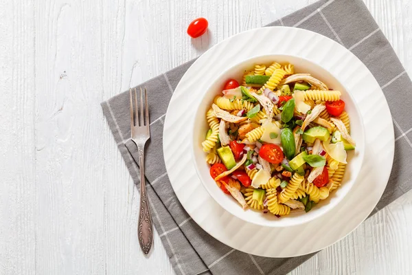 Healthy chicken pasta salad with avocado, tomato, olive oil and vinegar dressing in a white bowl on a white wooden table, horizontal top view, flat lay, copy space