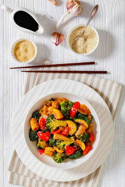 vegetable stir-fry of bell pepper, onion, zucchini, baby corn in cobs, broccoli florets poured with sticky soy sauce in white bowl on white wood table, vertical view from above