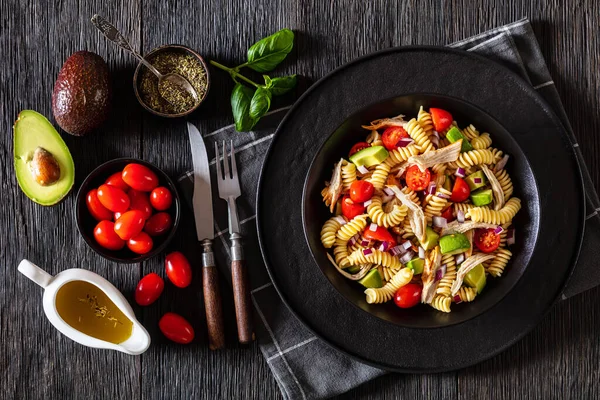 Healthy chicken pasta salad with avocado, tomato, olive oil and vinegar dressing in a black bowl on a dark wood table with ingredients, horizontal top view, flat lay