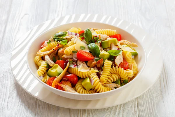 Healthy Chicken Pasta Salad with Avocado, Tomato, and olive oil and vinegar dressing in white bowl on white wood table,  horizontal view from above