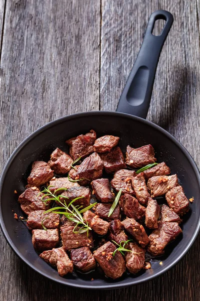 seared juicy new york strip steak bites with butter, rosemary and garlic on skillet on dark wood table, vertical view, copy space