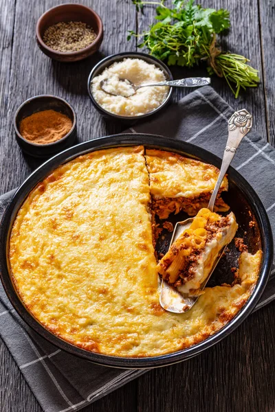 Greek Pastitsio of macaroni, ground lamb, grated cheese, and tomatoes topped with bechamel sauce and melted cheese in baking dish on dark wood table, vertical view