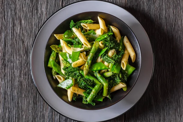 Pasta primavera with asparagus, snap peas, broccoli and kale in black bowl on dark wood table, horizontal view from above, flat lay, close-up