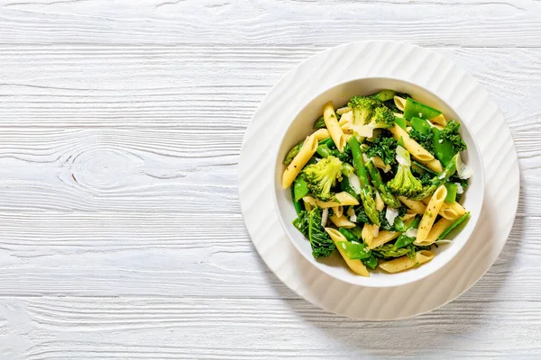 Pasta primavera with asparagus, snap peas, broccoli and kale in white bowl, on white wood table, horizontal view from above, flat lay, copy space