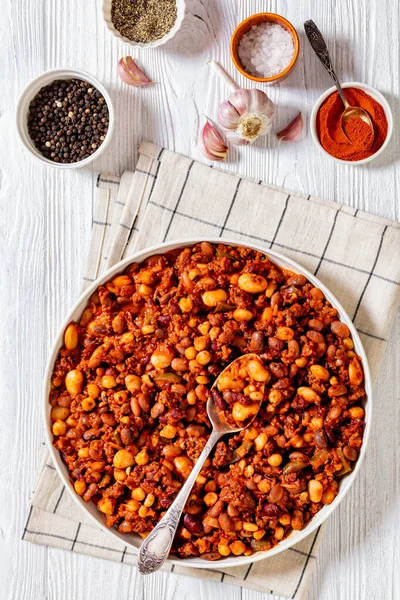 Beans and Sausage bake of kidney beans, great northern beans, black-eyed peas, pinto beans, chickpeas, meat and tomato sauce in baking dish on white wood table, vertical view from above