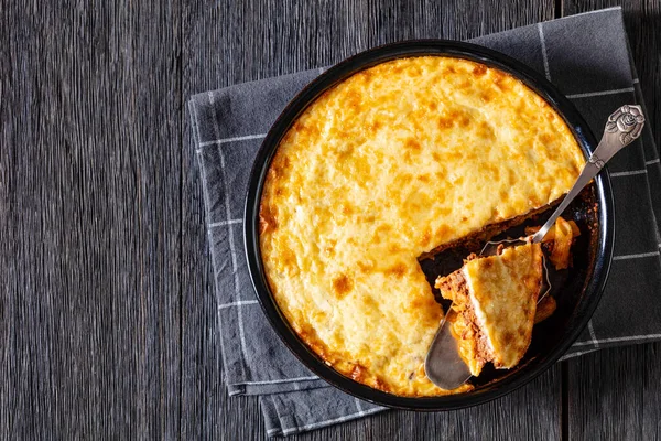 Greek Pastitsio of pasta, ground lamb, grated cheese and tomatoes topped with bechamel sauce and melted cheese in baking dish on dark wood table, horizontal view from above, flat lay, copy space