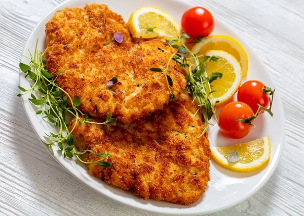 fried breaded chicken breast cutlets on white plate with lemon, fresh thyme and ripe tomatoes on white wood table, close-up