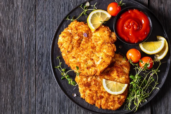 fried breaded chicken breast cutlets on black plate with tomato sauce, lemon, fresh thyme and ripe tomatoes on dark wood table, horizontal view from above, flat lay, free space, close-up