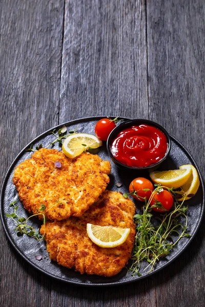 fried breaded chicken breast cutlets on black plate with tomato sauce, lemon, fresh thyme and ripe tomatoes on dark wood table, vertical view, copy space