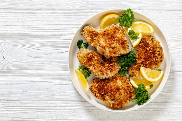 baked in oven panko breaded pork chops on bone with lemon slices and fresh parsley on plate on white textured wood table, horizontal view from above, flat lay, free space