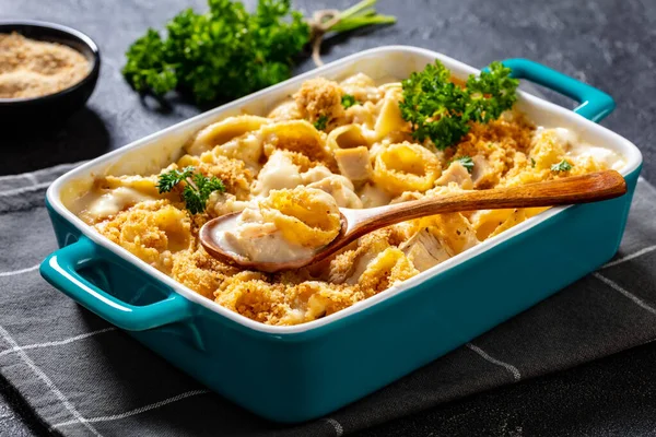 close-up of cheesy and creamy chicken and shells pasta bake in baking dish on dark grey concrete table, horizontal view from above