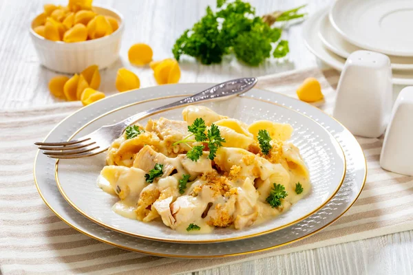 cheesy and creamy chicken and shells pasta bake in white bowl with fork on white table, horizontal view from above, close-up